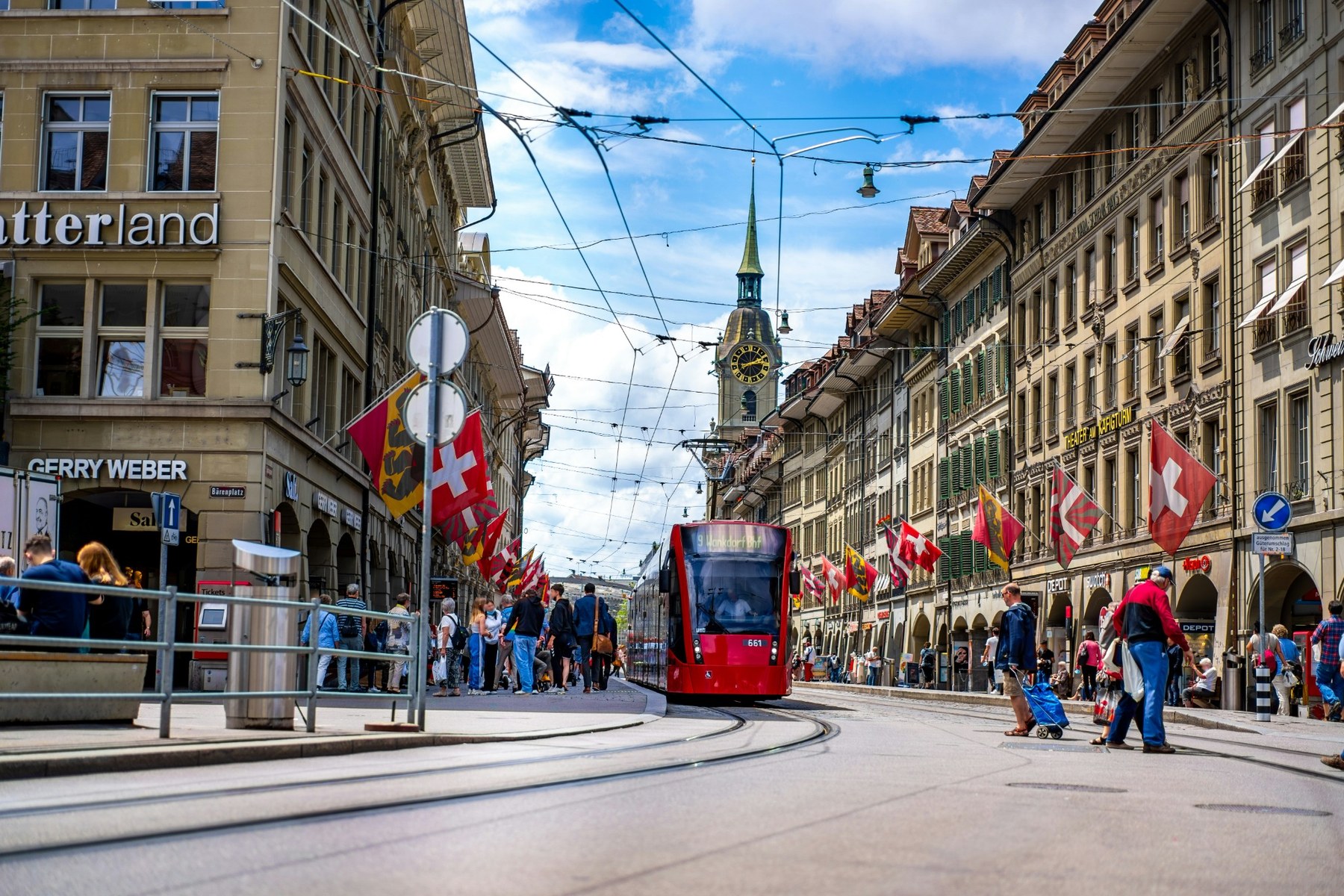 A tram moving through a busy Bern street among pedestrians and Swiss flags.